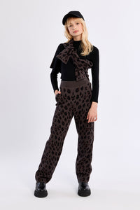 A model stands against a light gray studio background wearing a black long-sleeve top with a large dark brown bow, dark brown leopard-print pants, and a black beret.
