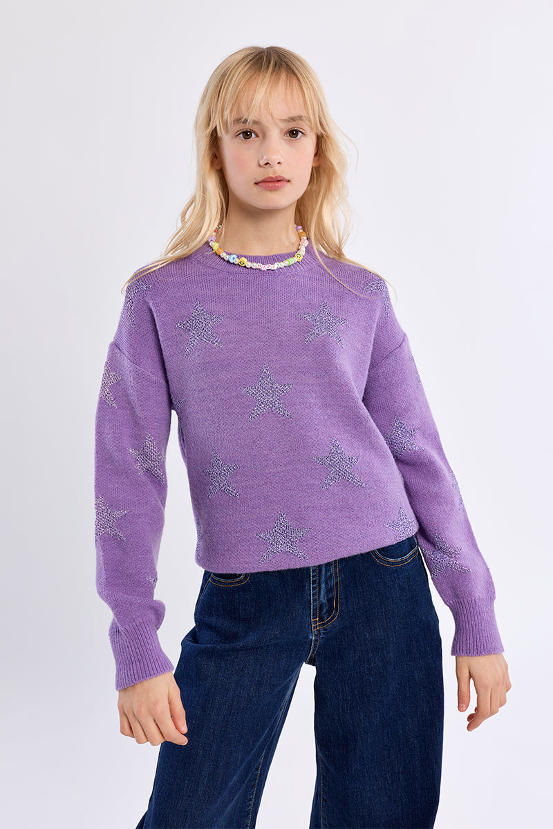 A young blonde girl wearing a lavender purple knit sweater with silver glittery star patterns and a colorful beaded collar, paired with dark blue jeans, against a plain light gray background.