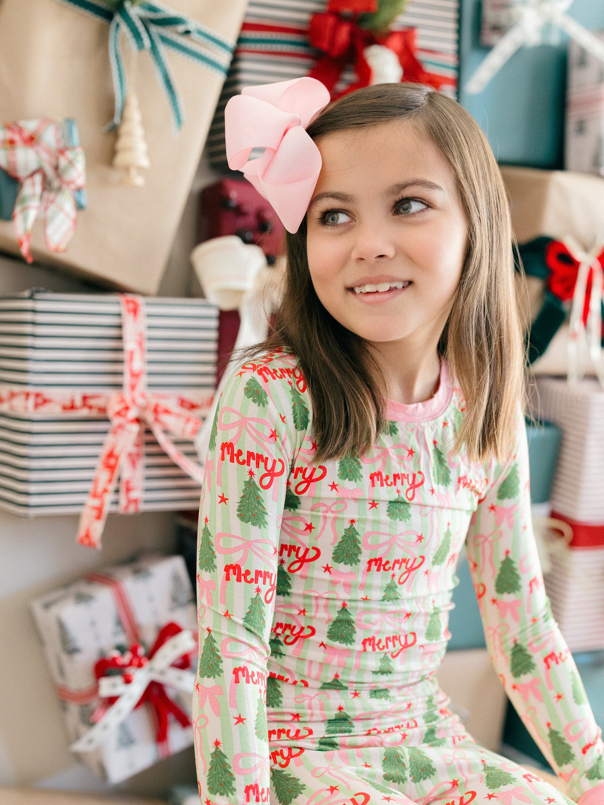 A young girl with a pink bow in her hair looks to the side while wearing a festive long-sleeve pajama top with a pink and green plaid pattern, decorated with red 'Merry' text and green Christmas trees. She is surrounded by wrapped holiday presents.