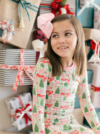 A young girl with a pink bow in her hair looks to the side while wearing a festive long-sleeve pajama top with a pink and green plaid pattern, decorated with red 'Merry' text and green Christmas trees. She is surrounded by wrapped holiday presents.