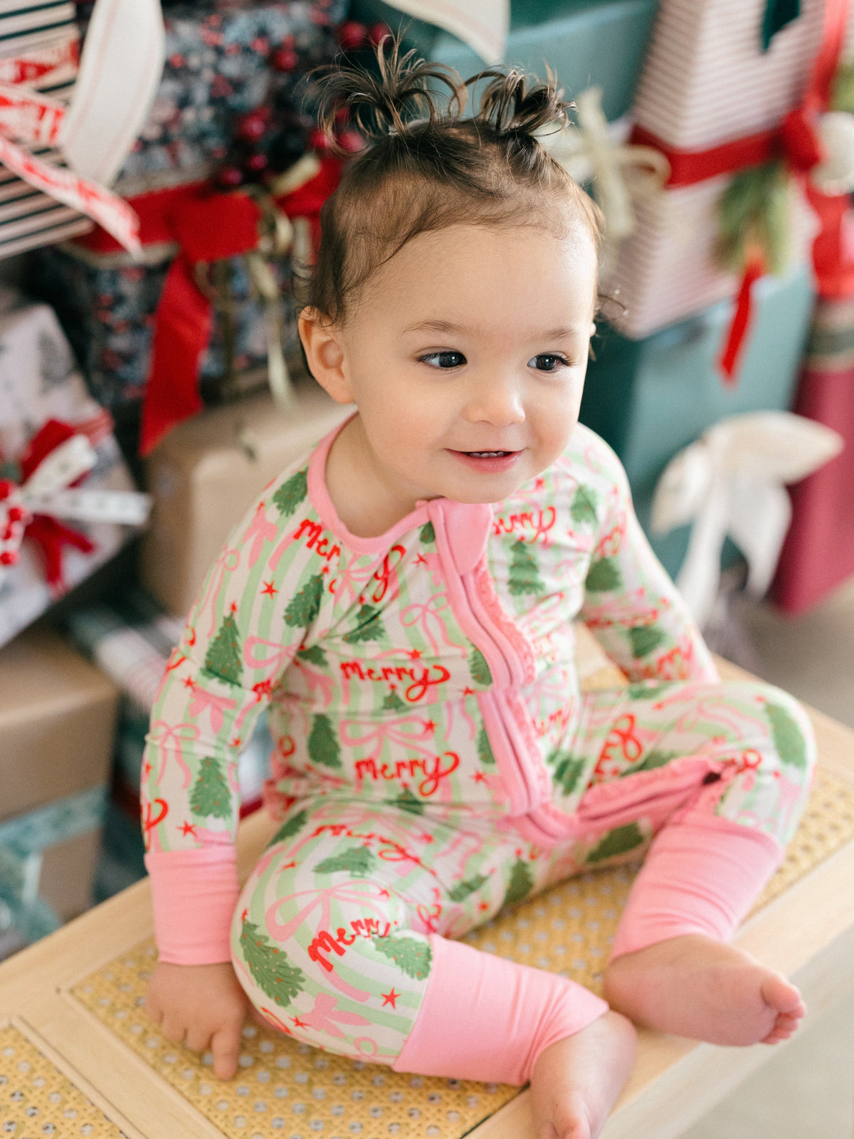 A baby sitting on a wooden surface wearing a festive Christmas-themed footed sleeper. The sleeper has a light pink background with green Christmas trees, red 'Merry' text, and pink trim and leg cuffs.