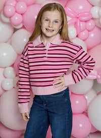 A young girl with blonde hair stands in front of a pink and white balloon wall, wearing a pink and dark red striped knit polo sweater with blue jeans.