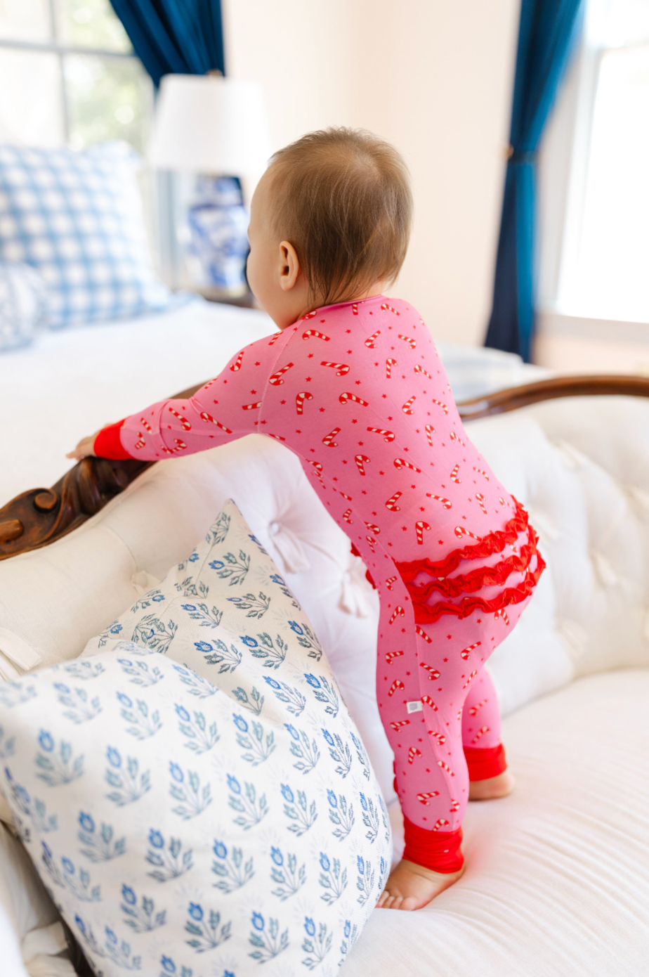 A baby wearing pink candy cane print pajamas stands on a white tufted sofa in a bright bedroom, holding onto a wooden headboard.