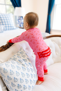 A baby wearing pink candy cane print pajamas stands on a white tufted sofa in a bright bedroom, holding onto a wooden headboard.