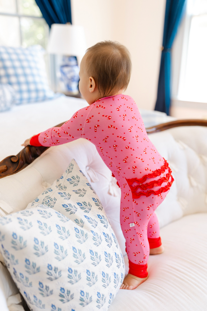 A baby wearing pink candy cane print pajamas stands on a white tufted sofa in a bright bedroom, holding onto a wooden headboard.