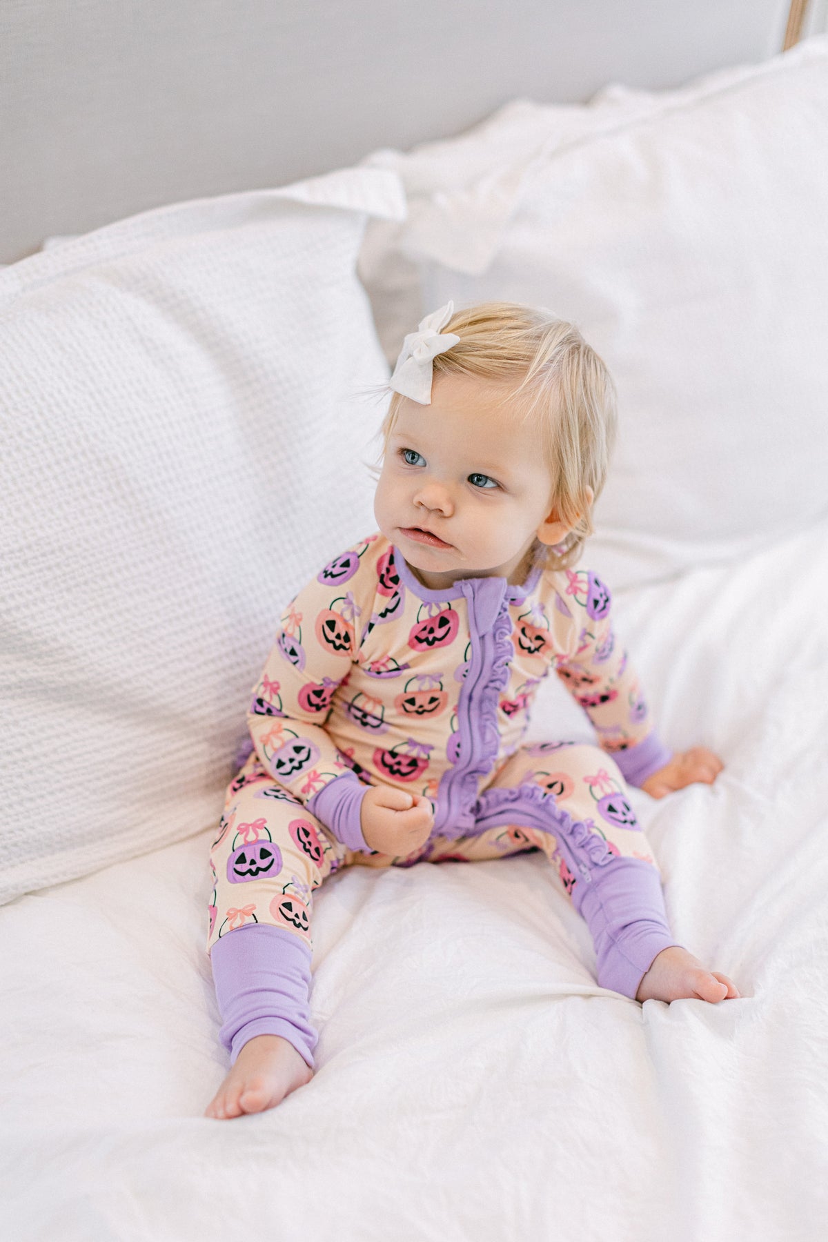 A toddler girl with blonde hair and a white bow sits on a white bed wearing a light pink and purple onesie pajama with a cartoon character print.