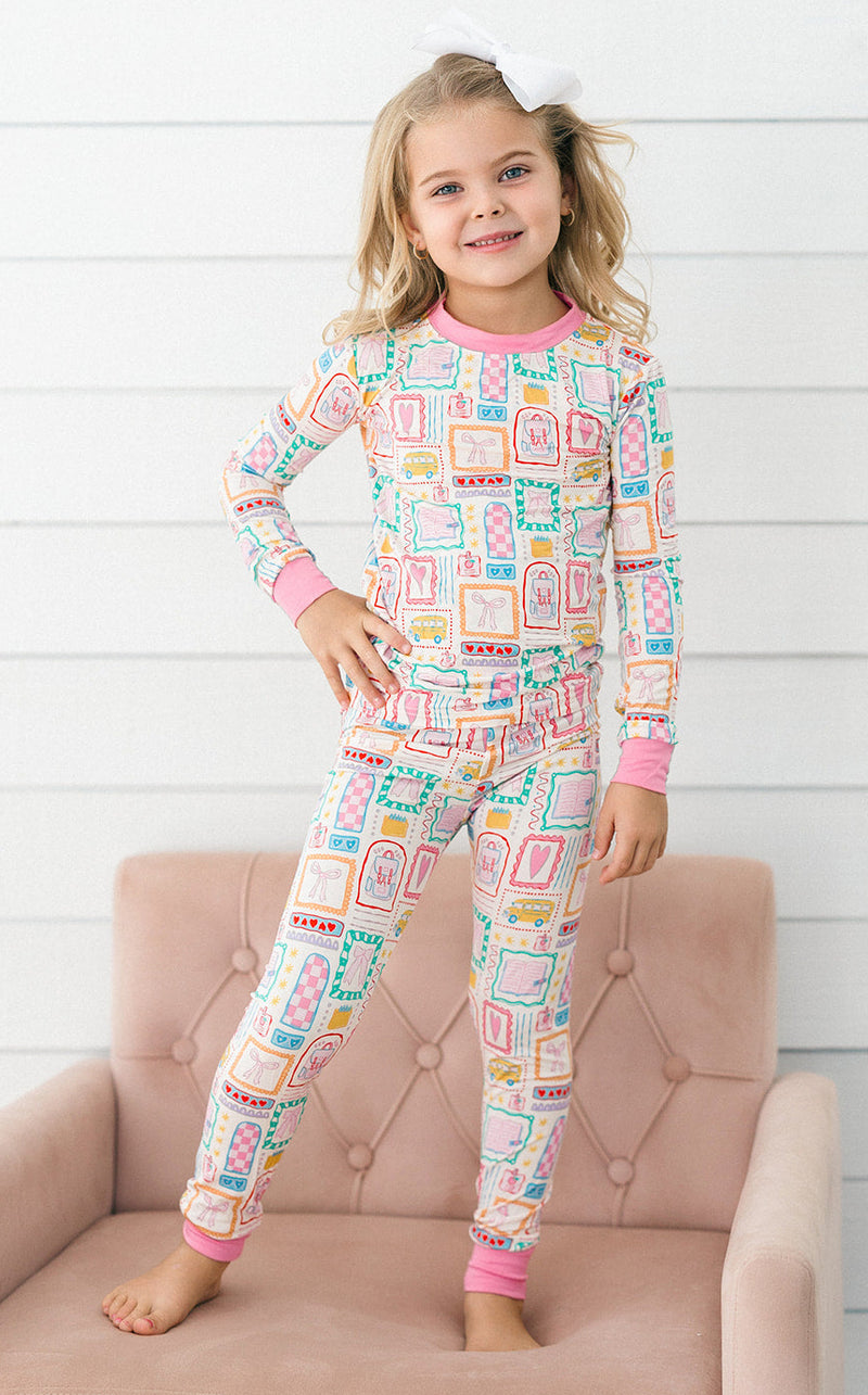 A young girl stands barefoot on a pink tufted chair against a white wood-paneled wall, wearing a long-sleeve pajama top and matching pajama pants with a colorful geometric pattern and pink trim.