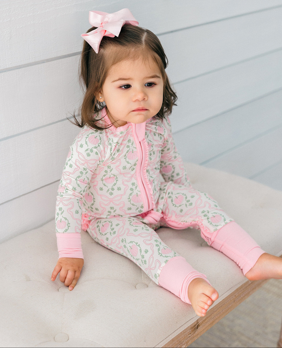 A toddler girl wearing a pink and white paisley print zip-up sleeper sits on a tufted bench against a white paneled wall.