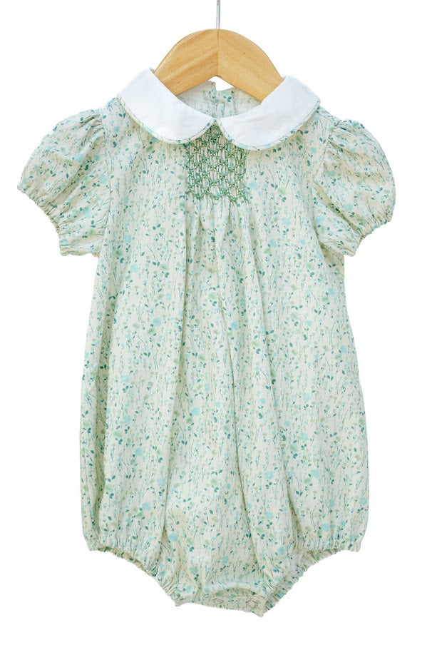 A baby romper in a light sage green and white floral print, featuring a white Peter Pan collar and a smocked chest detail, hanging on a wooden hanger against a white background.
