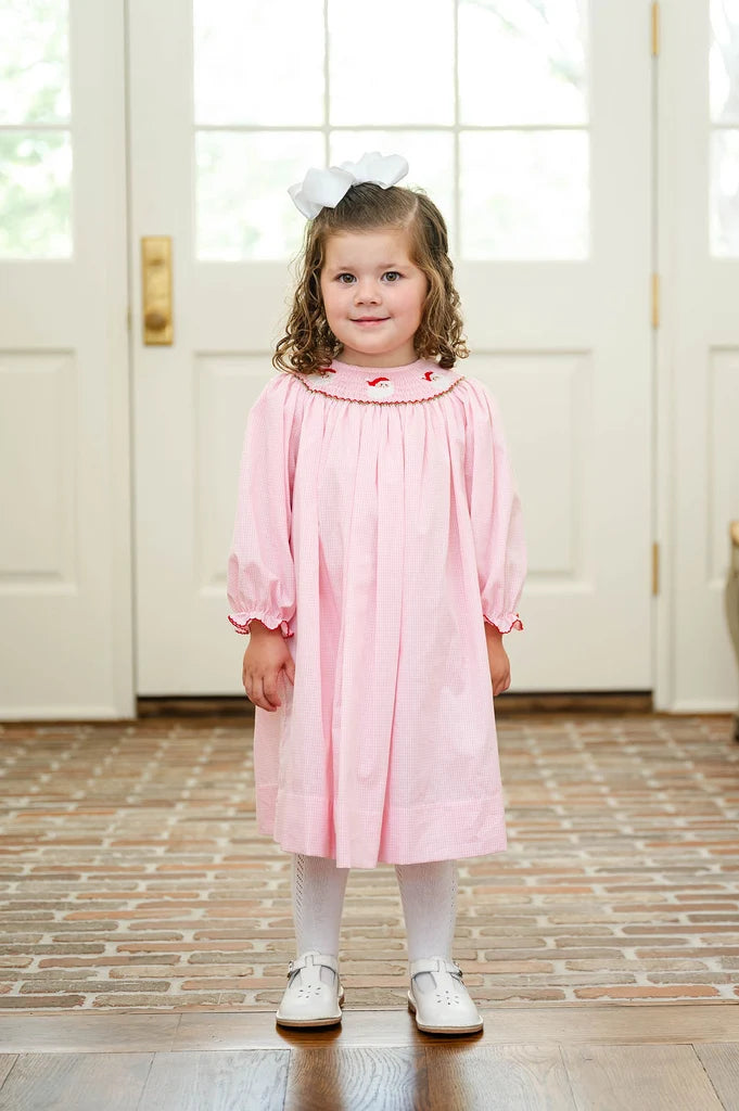 A young girl with curly brown hair wearing a light pink smocked dress, white tights, and white shoes, standing in front of white French doors with a brick floor.