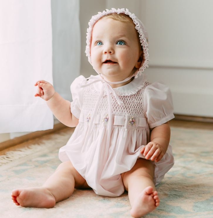 A baby girl wearing a light pink smocked romper and matching bonnet sits on a patterned rug in a bright indoor room.