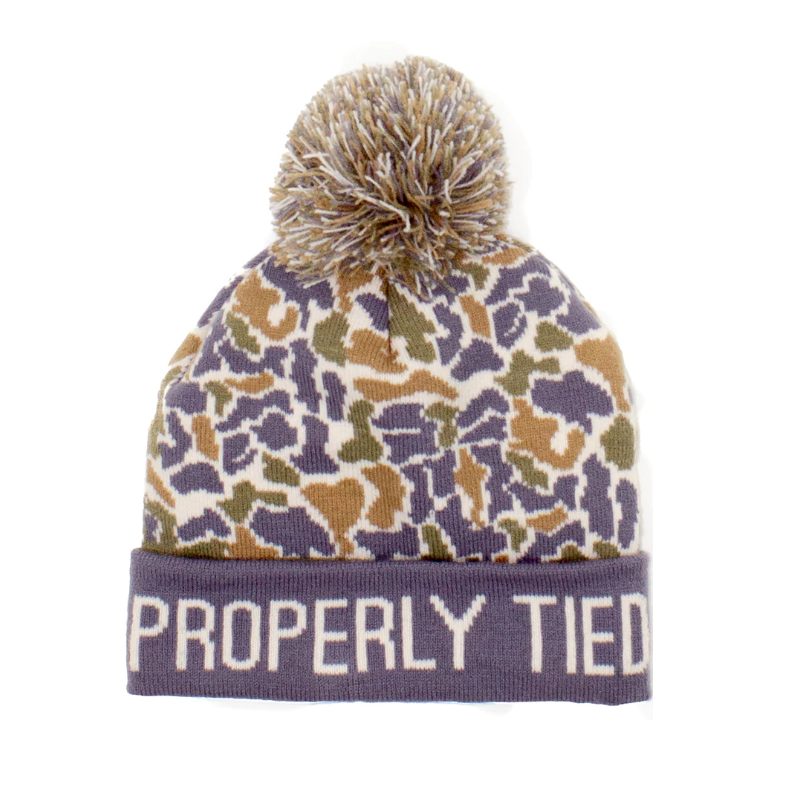 A knit pom-pom beanie hat featuring a purple, beige, and olive green camouflage pattern on the upper portion, with a solid purple ribbed cuff displaying the text 'PROPERLY TIED' in white. It is photographed against a plain white background.