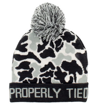 A knit pom-pom beanie hat featuring a black, gray, and off-white camouflage pattern, with a black cuff displaying the white text 'PROPERLY TIED' and a gray and black pom-pom on top.