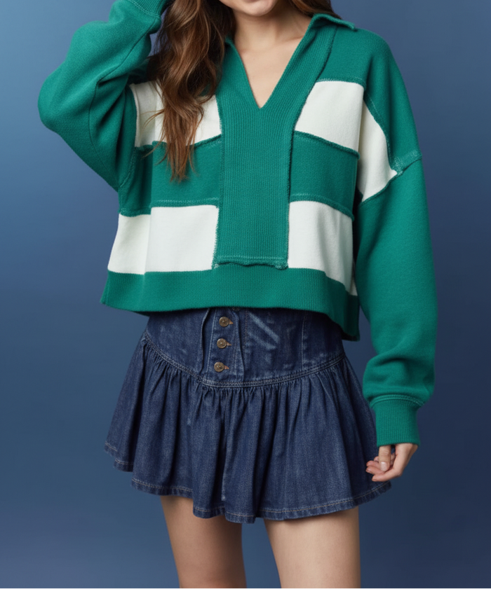 A young woman wearing a green and white color-block knit cardigan sweater and a dark blue denim pleated mini skirt, posing against a textured blue studio background.
