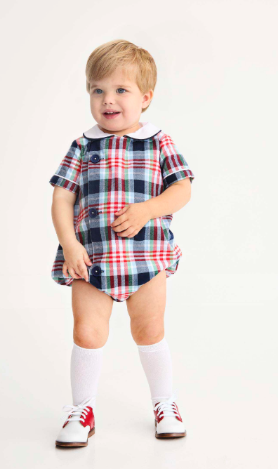 A toddler standing against a plain white studio background, wearing a short-sleeve plaid romper in navy, red, white, and light blue with a white collar, white knee-high socks, and white shoes with red accents.