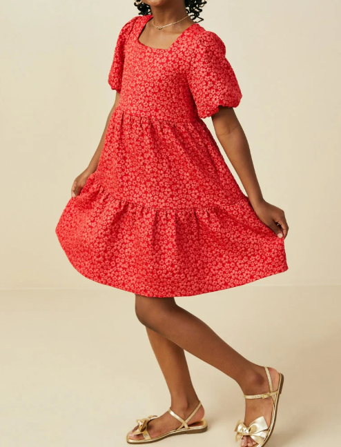 A woman modeling a red short-sleeve tiered dress with a small white floral print, paired with gold sandals, against a solid beige studio background.