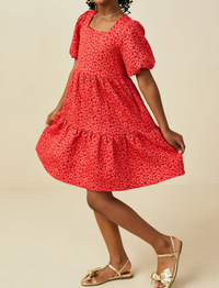 A woman modeling a red short-sleeve tiered dress with a small white floral print, paired with gold sandals, against a solid beige studio background.