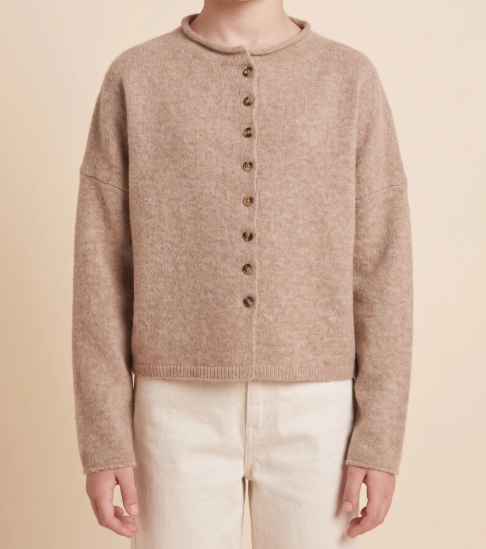 A person wearing a cropped, long-sleeve knit cardigan in a warm taupe-beige color with a row of dark brown buttons down the front, paired with cream pants, against a solid beige background.