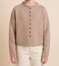 A person wearing a cropped, long-sleeve knit cardigan in a warm taupe-beige color with a row of dark brown buttons down the front, paired with cream pants, against a solid beige background.