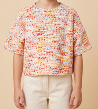 A child wearing a short-sleeve textured knit top with a multicolor marled pattern of orange, yellow, red, and blue on a white base, paired with cream pants, against a solid beige background.