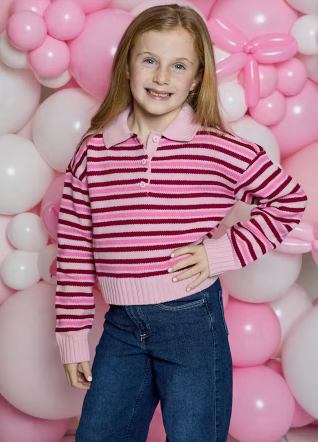 A young girl with blonde hair stands in front of a pink and white balloon wall, wearing a pink and dark red striped knit polo sweater with blue jeans.