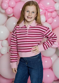 A young girl with blonde hair stands in front of a pink and white balloon wall, wearing a pink and dark red striped knit polo sweater with blue jeans.