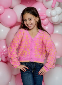 A young girl with long brown hair smiling, wearing a fuzzy pink cardigan with yellow-orange leopard spots and blue jeans, standing in front of a pink and white balloon backdrop.