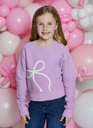 A young girl with light brown hair stands smiling in front of a pink and white balloon backdrop, wearing a lavender ribbed knit sweater with a white bow design on the front and blue jeans.
