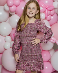 A young girl with long blonde hair stands in front of a pink and white balloon backdrop, wearing a long-sleeve tiered dress with a pink, purple, and white floral pattern.