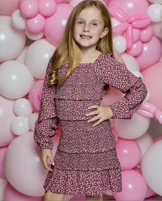 A young girl with long blonde hair stands in front of a pink and white balloon backdrop, wearing a long-sleeve tiered dress with a pink, purple, and white floral pattern.