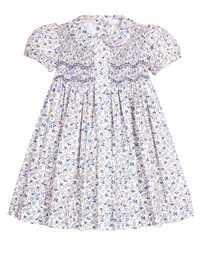 A short-sleeve baby dress with a white base, blue and light pink floral print, smocked bodice, and Peter Pan collar, shown against a plain white background.