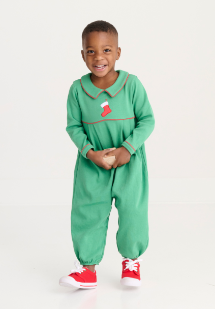 A young child wearing a festive green long-sleeve footless jumpsuit with a red trim and a small red stocking graphic on the chest, paired with red sneakers, standing against a plain white studio background.