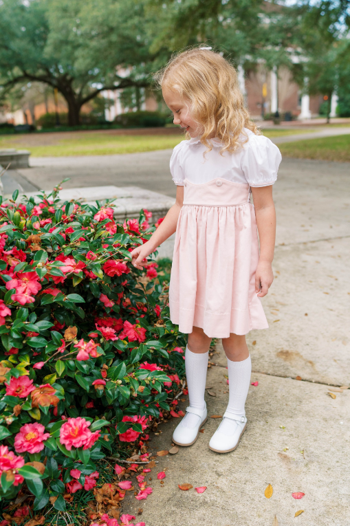 A young girl with blonde curly hair stands on a concrete sidewalk next to a bush of red flowers, wearing a light pink and white midi dress with puffed sleeves, white knee-high socks, and white shoes.