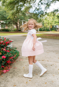 A young girl with blonde hair is smiling and posing outdoors on a concrete path, wearing a light pink and white dress with white knee-high socks and white shoes.