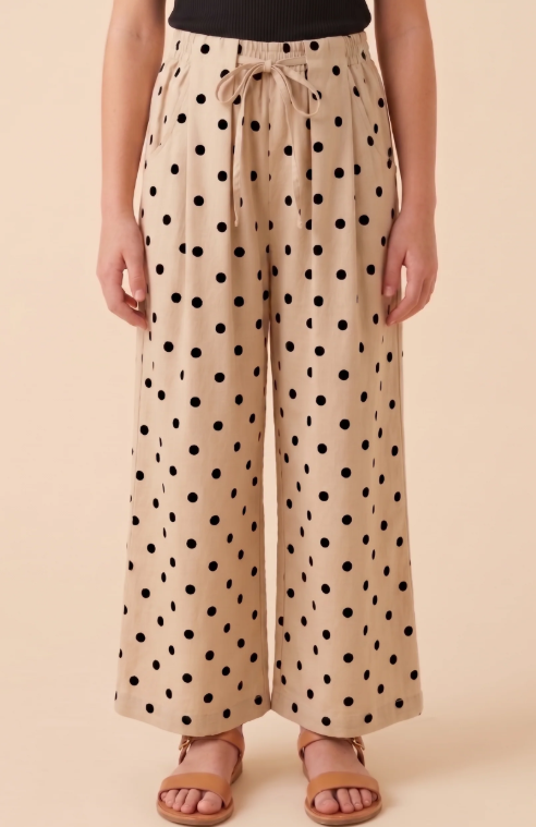 A person wearing beige wide-leg drawstring pants with a black polka dot pattern, paired with a black top and tan slide sandals, against a solid beige studio background.