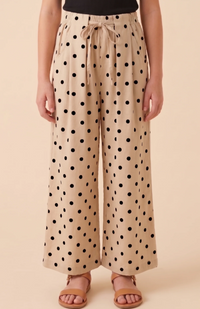 A person wearing beige wide-leg drawstring pants with a black polka dot pattern, paired with a black top and tan slide sandals, against a solid beige studio background.