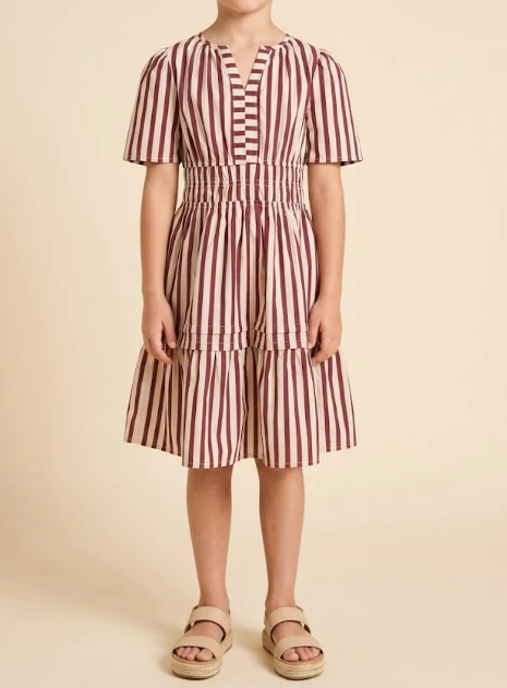 A child wearing a short-sleeve tiered dress with vertical burgundy and cream stripes, paired with beige platform sandals, standing against a solid beige background.