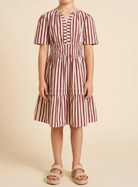 A child wearing a short-sleeve tiered dress with vertical burgundy and cream stripes, paired with beige platform sandals, standing against a solid beige background.