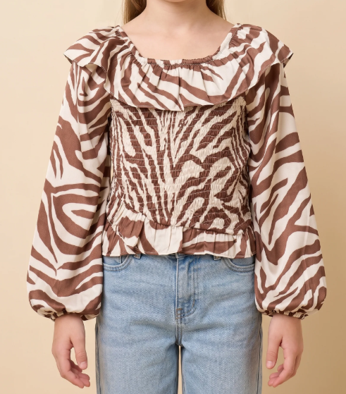 A child wearing a brown and cream zebra-print blouse with a smocked bodice and ruffled neckline, paired with light blue jeans, against a solid beige background.