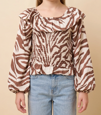 A child wearing a brown and cream zebra-print blouse with a smocked bodice and ruffled neckline, paired with light blue jeans, against a solid beige background.
