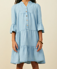 A light blue tiered cotton dress with a button-down front and ruffled sleeves, shown on a model against a plain beige background.