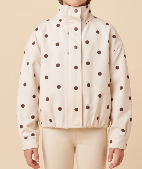 A model wearing a cream-colored jacket with dark brown polka dots, paired with matching cream pants, against a solid beige background.