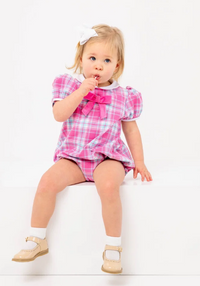 A toddler girl with blonde hair and a white bow sits on a white step, wearing a pink and white plaid romper with a large pink bow at the collar, white socks, and tan Mary Jane shoes.