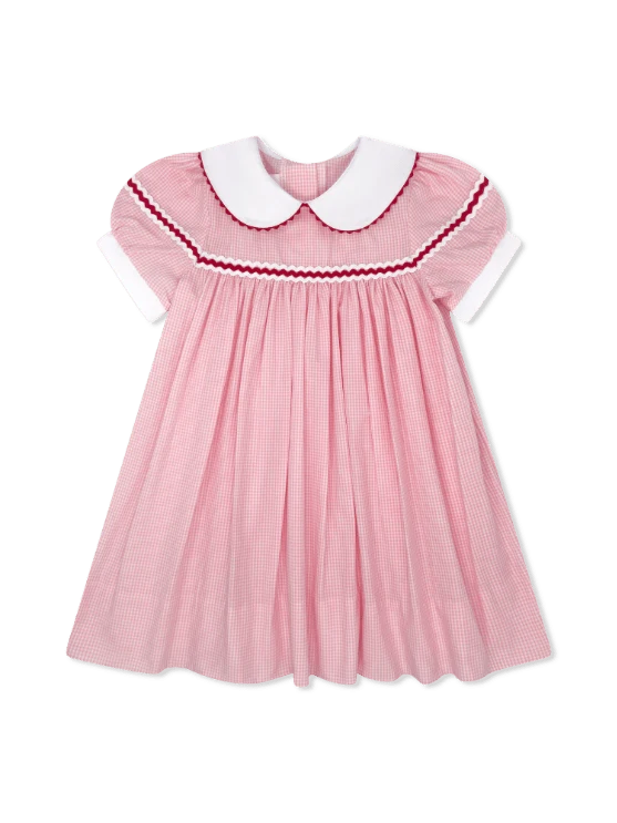 A pink gingham baby dress with a white Peter Pan collar, white short sleeves, and red and white smocked trim around the neckline.