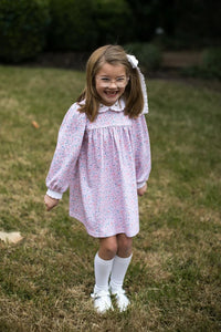 A young girl with glasses and a white hair bow stands outdoors on a grassy lawn, wearing a light pink long-sleeve dress with a small blue and yellow floral print, white knee-high socks, and white shoes.