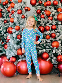 A young girl stands barefoot in front of decorated Christmas trees, wearing a blue long-sleeve pajama set with a green and white pine tree pattern and a green bow in her hair.