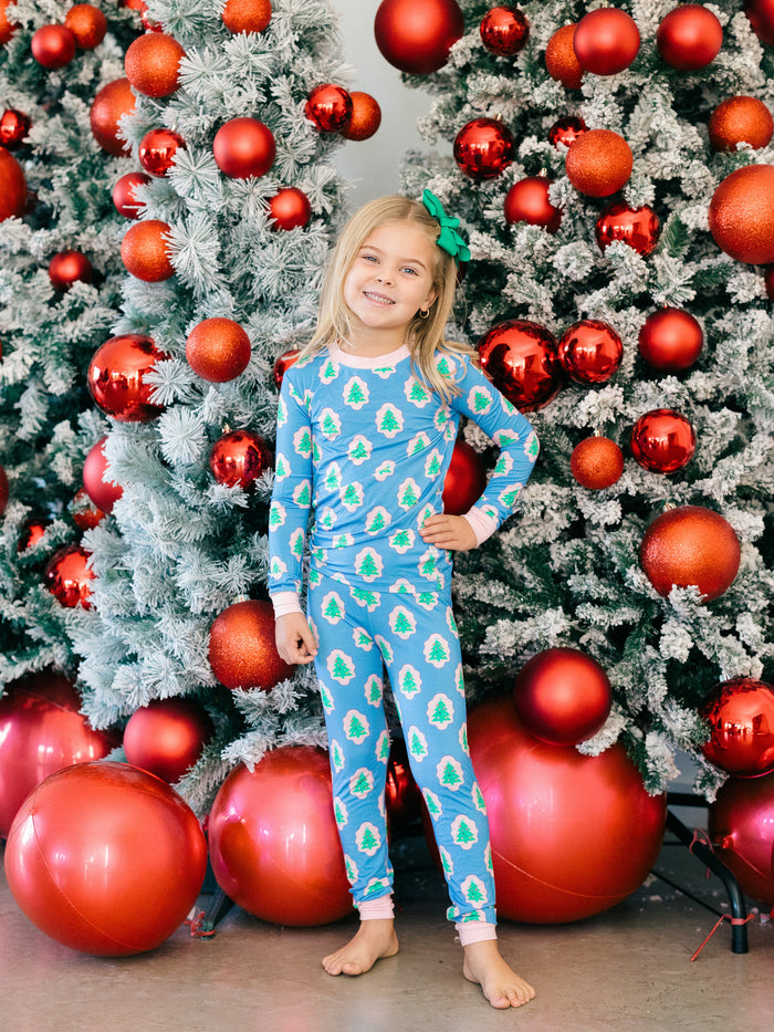 A young girl stands barefoot in front of decorated Christmas trees, wearing a blue long-sleeve pajama set with a green and white pine tree pattern and a green bow in her hair.