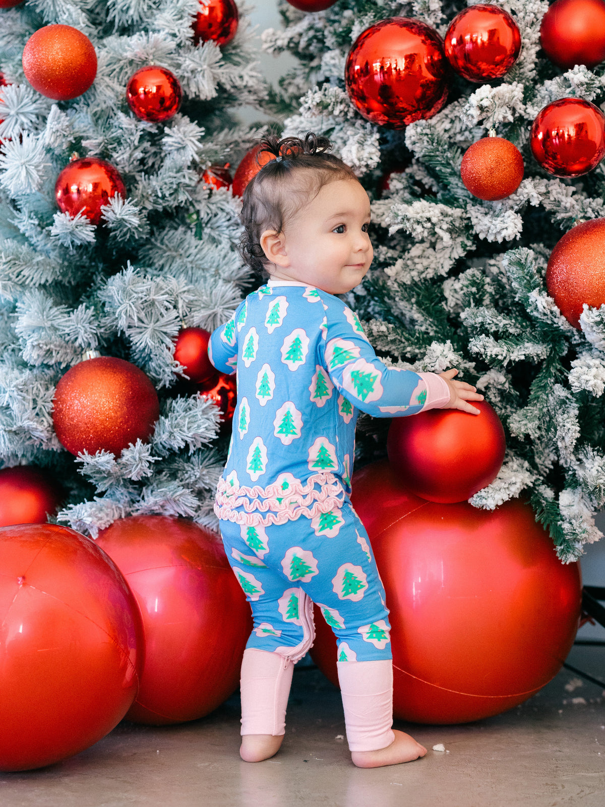 A toddler stands barefoot in front of a decorated Christmas tree, wearing a light blue one-piece pajama with a green pine tree pattern and pink trim, paired with light pink leggings.