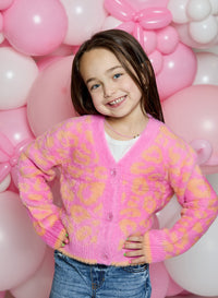 A young girl smiling in front of a pink and white balloon backdrop, wearing a fuzzy pink cardigan with yellow-orange leopard-like spots over a white shirt and blue jeans.