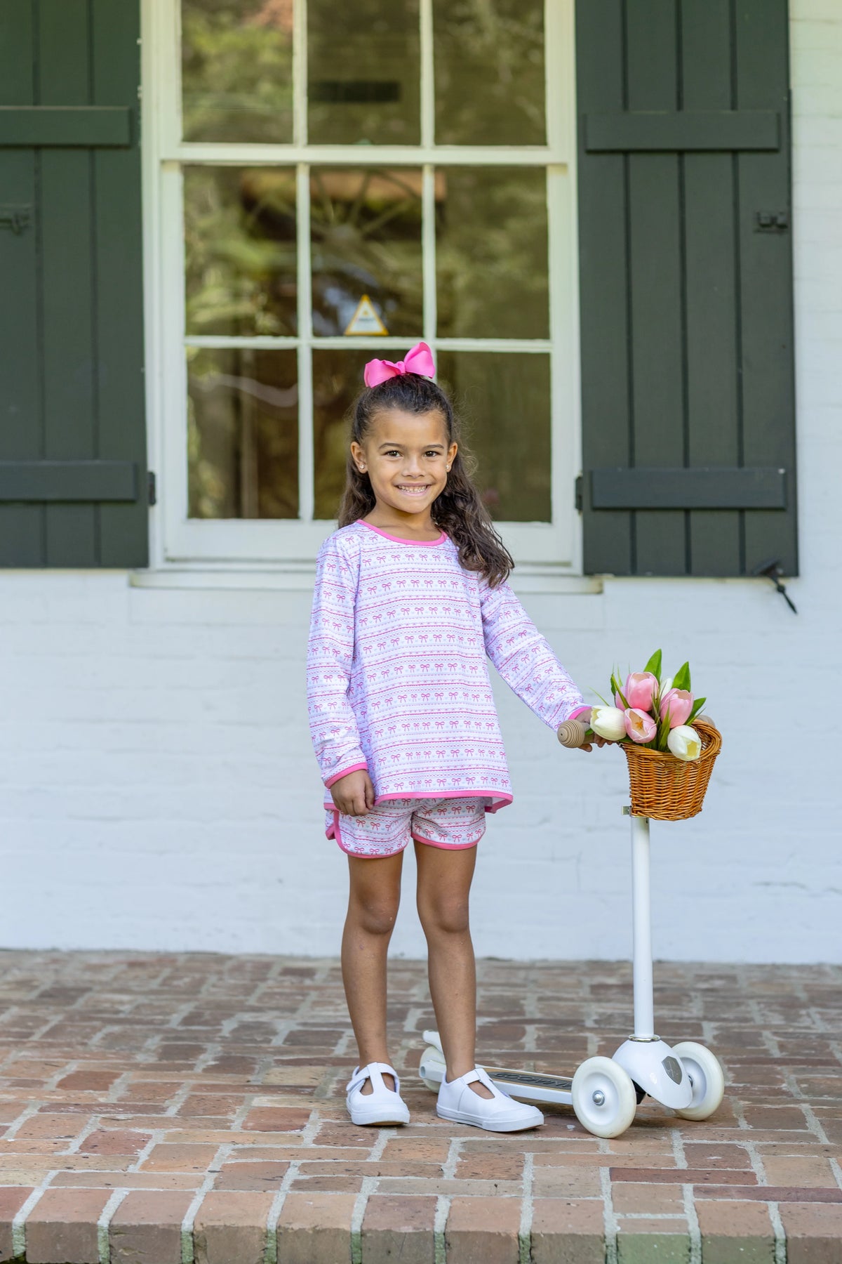 A young girl stands on a brick porch in front of a white wall with dark green shutters, wearing a light pink and white patterned long-sleeve shirt and matching shorts, holding a small basket of flowers next to a white scooter.